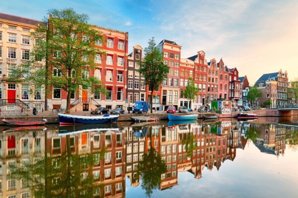 A picturesque canal in Amsterdam with colorful houses lining the water's edge. Boats are moored along the canal, and a bridge crosses the waterway in the background. The sky is blue with some clouds.