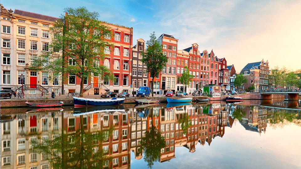 A picturesque canal in Amsterdam with colorful houses lining the water's edge. Boats are moored along the canal, and a bridge crosses the waterway in the background. The sky is blue with some clouds.