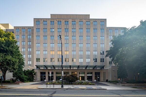 Front view of The Brookings Institution building with a symmetrical facade, glass canopy entrance, landscaped greenery, and trees on a sunny day.