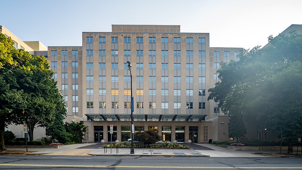 Front view of The Brookings Institution building with a symmetrical facade, glass canopy entrance, landscaped greenery, and trees on a sunny day.