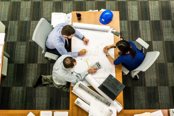 Three individuals collaborate at a table, examining blueprints and papers for a project discussion.