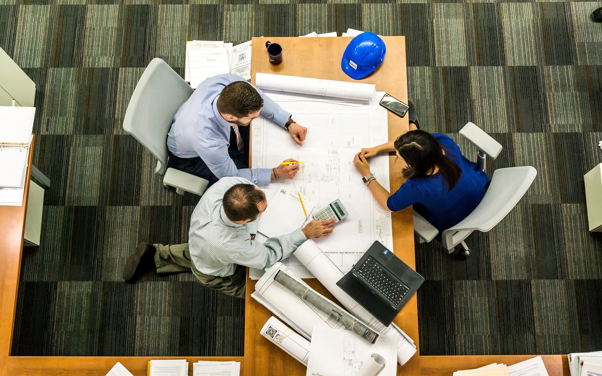 Three individuals collaborate at a table, examining blueprints and papers for a project discussion.