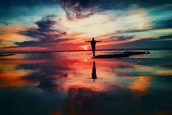 Silhouette of a person with arms outstretched on reflective water during a vibrant sunset with orange, pink, and blue hues.