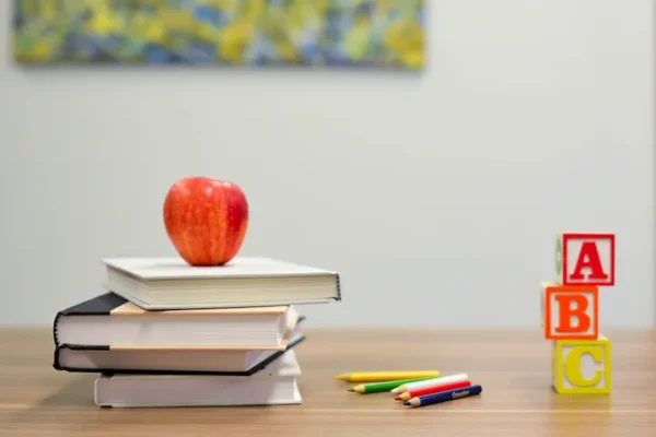 Stack of books with a red apple on top, colorful pencils, and ABC blocks on a wooden desk.