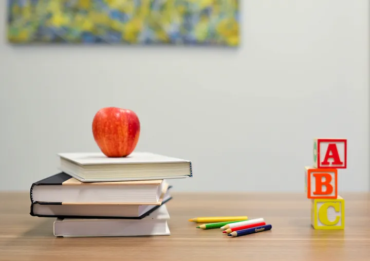 Stack of books with a red apple on top, colorful pencils, and ABC blocks on a wooden desk.